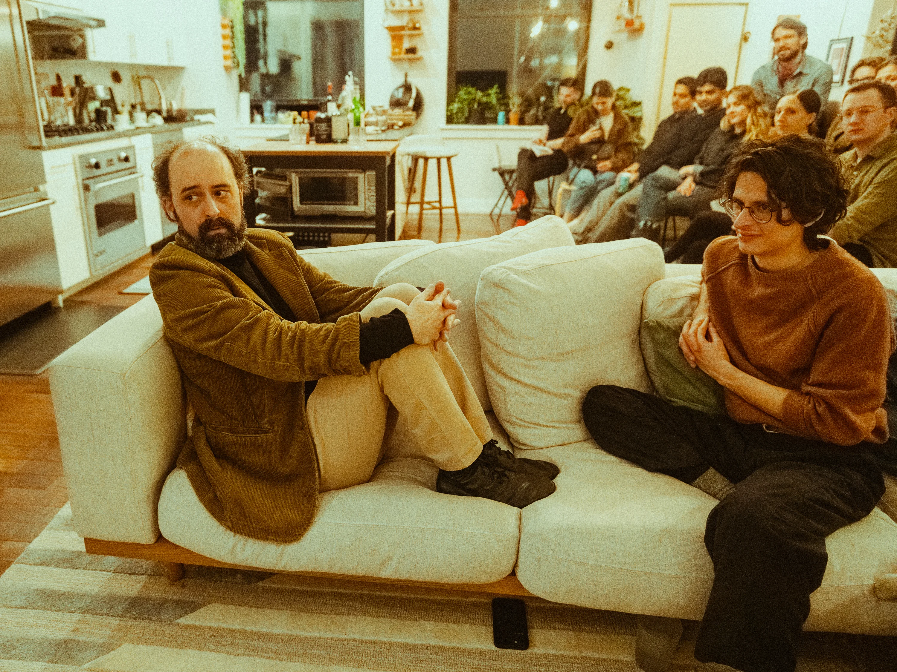 Two actors seated closely on a sofa during a performance in a private home